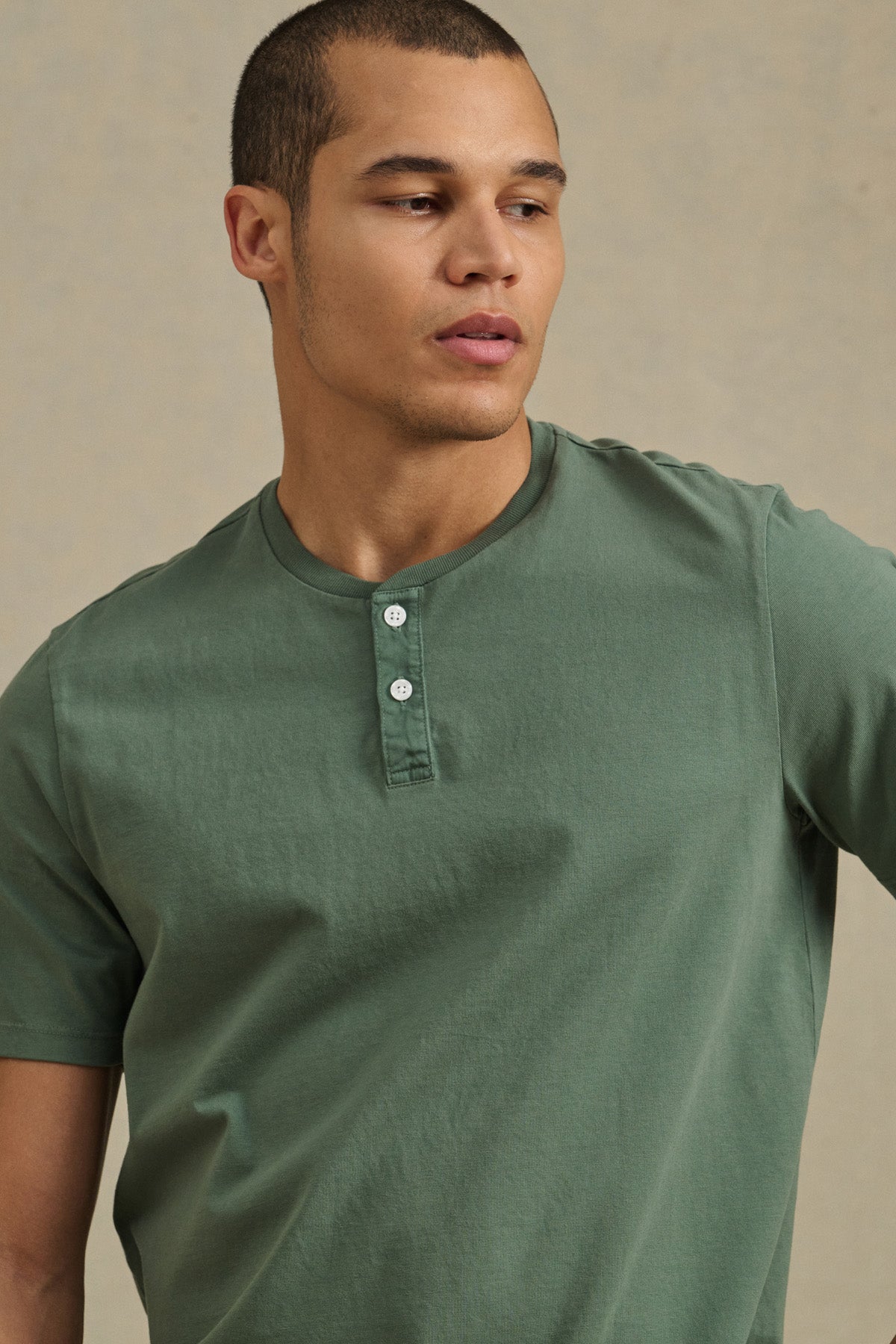 A person wearing the Velvet by Graham & Spencer FELIX HENLEY—a green short sleeve, three-button top—looks to the side against a neutral background.-41479917174977