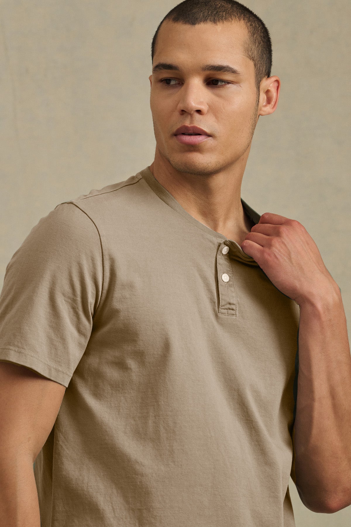 A man wearing the FELIX HENLEY by Velvet by Graham & Spencer looks to the side, touching the collar near two buttons of his beige short sleeve jersey shirt.-41435170341057