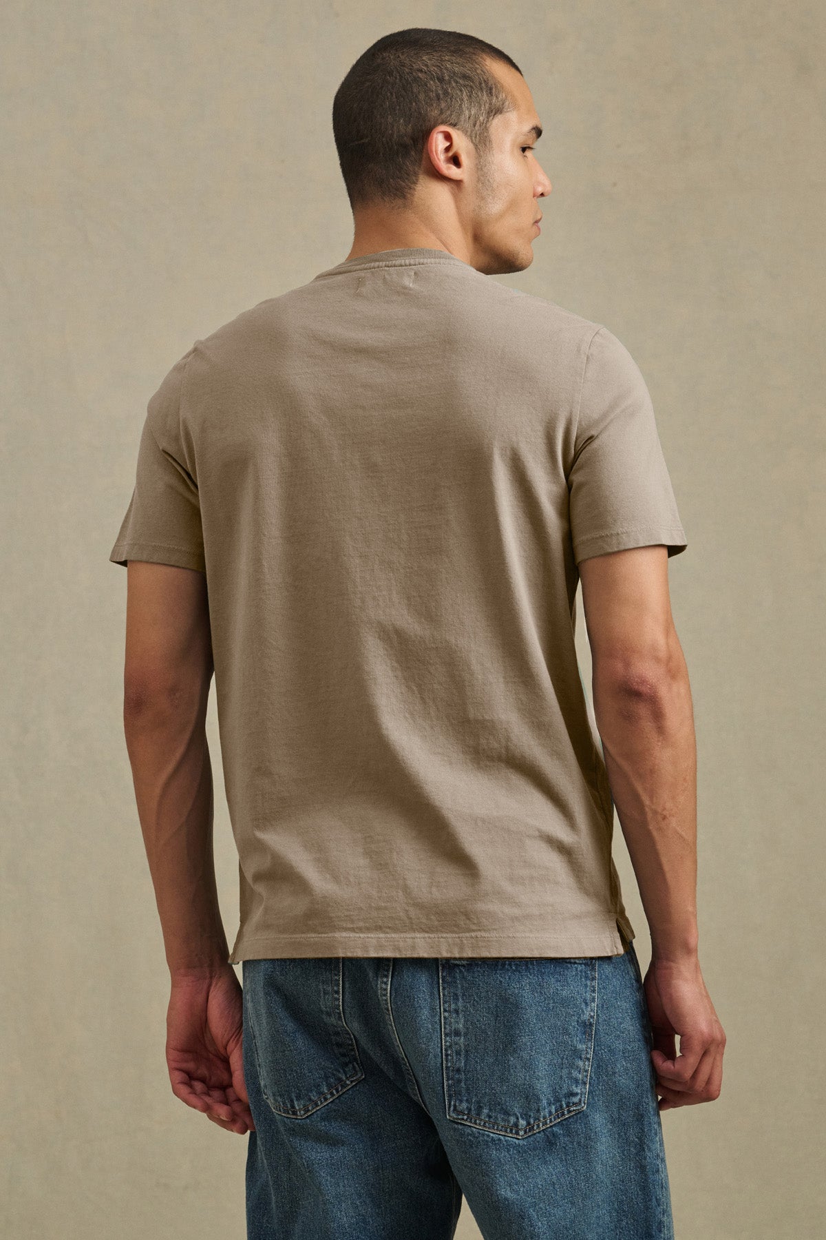 A man stands facing away from the camera against a neutral background, wearing the FELIX HENLEY by Velvet by Graham & Spencer in beige with blue jeans.