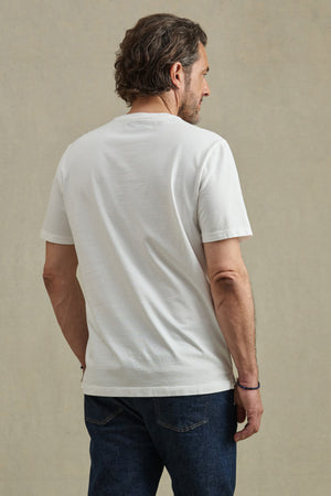 A man with short brown hair and a beard faces away from the camera, wearing a plain white Velvet by Graham & Spencer FELIX HENLEY shirt and dark blue jeans against a beige background.