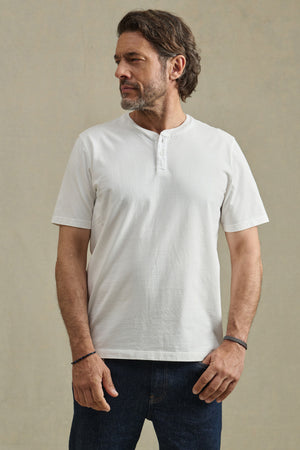 Man with short brown hair and beard wears the Velvet by Graham & Spencer FELIX HENLEY in white with dark jeans, standing against a plain beige background.