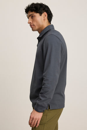 A man stands in profile wearing the FLINT POLO by Velvet by Graham & Spencer, a classic long-sleeve dark gray collared shirt with ribbed cuffs, paired with olive green pants against a plain light background.