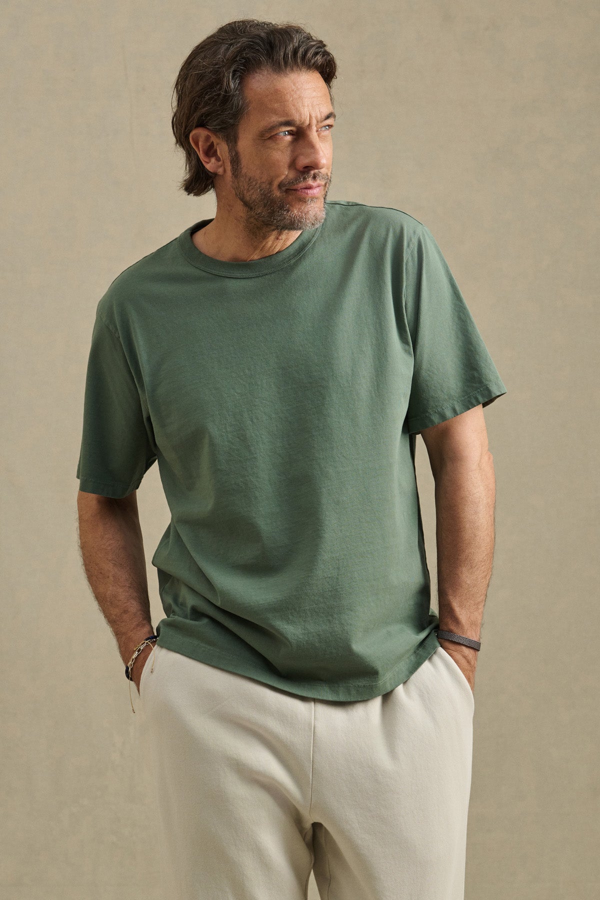 Man with medium-length hair and beard wears the WELLS TEE by Velvet by Graham & Spencer, a relaxed fit green Pima cotton jersey t-shirt, paired with light pants, hands in pockets against a plain background.