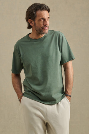 Man with medium-length hair and beard wears the WELLS TEE by Velvet by Graham & Spencer, a relaxed fit green Pima cotton jersey t-shirt, paired with light pants, hands in pockets against a plain background.
