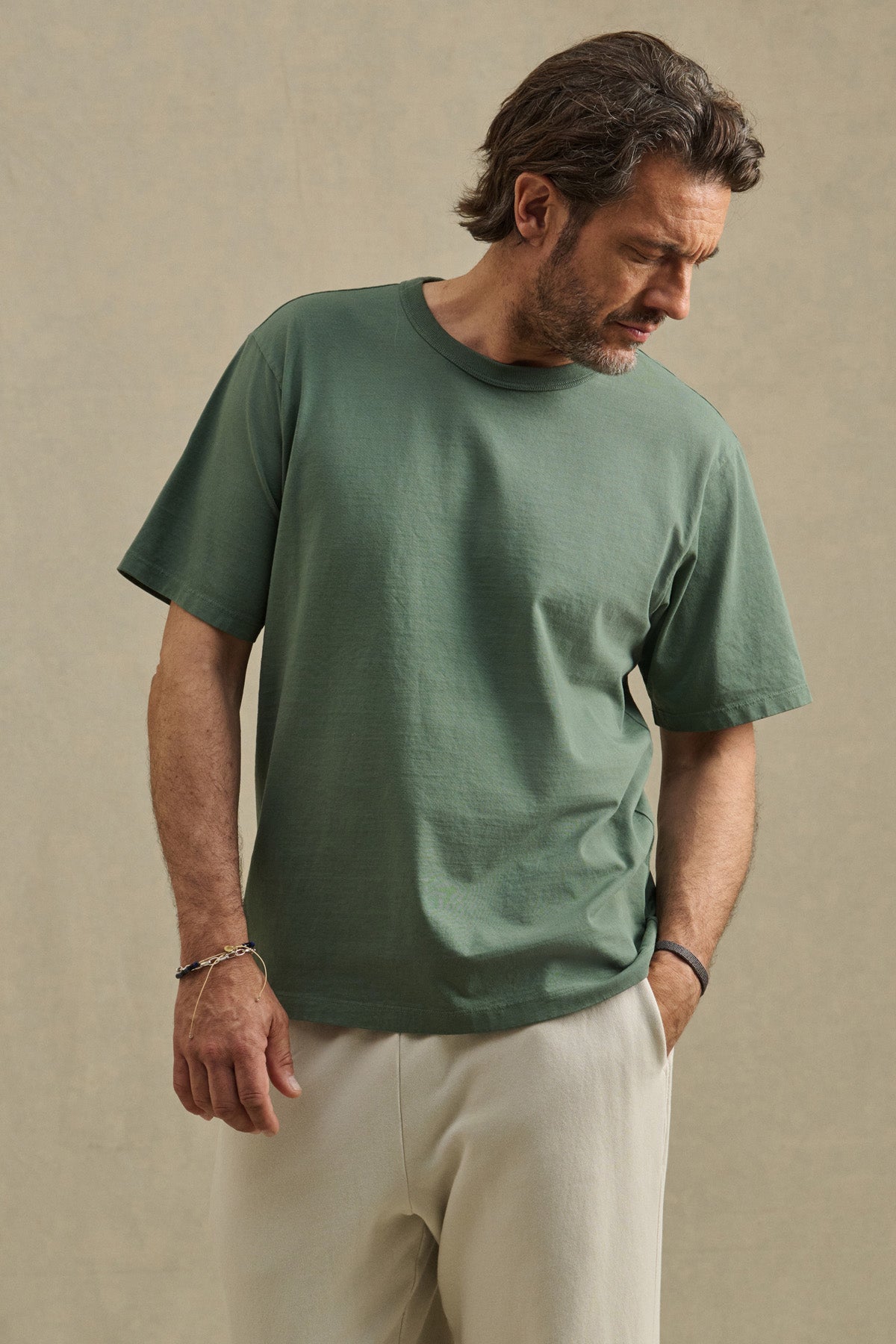 Wearing the WELLS TEE by Velvet by Graham & Spencer in green Pima cotton and light beige pants, a man stands against a plain background, looking down to his left—showcasing a relaxed fit perfect for all-day comfort.-41480101429441