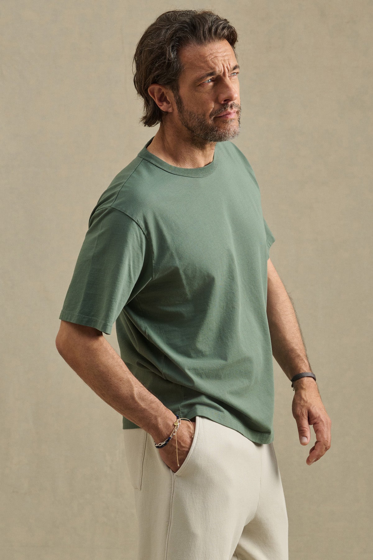 A man stands against a neutral background, wearing the Velvet by Graham & Spencer WELLS TEE in green—a relaxed fit Pima cotton jersey—paired with light-colored pants, one hand in his pocket as he looks slightly upward.-41480101462209
