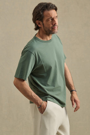 A man stands against a neutral background, wearing the Velvet by Graham & Spencer WELLS TEE in green—a relaxed fit Pima cotton jersey—paired with light-colored pants, one hand in his pocket as he looks slightly upward.