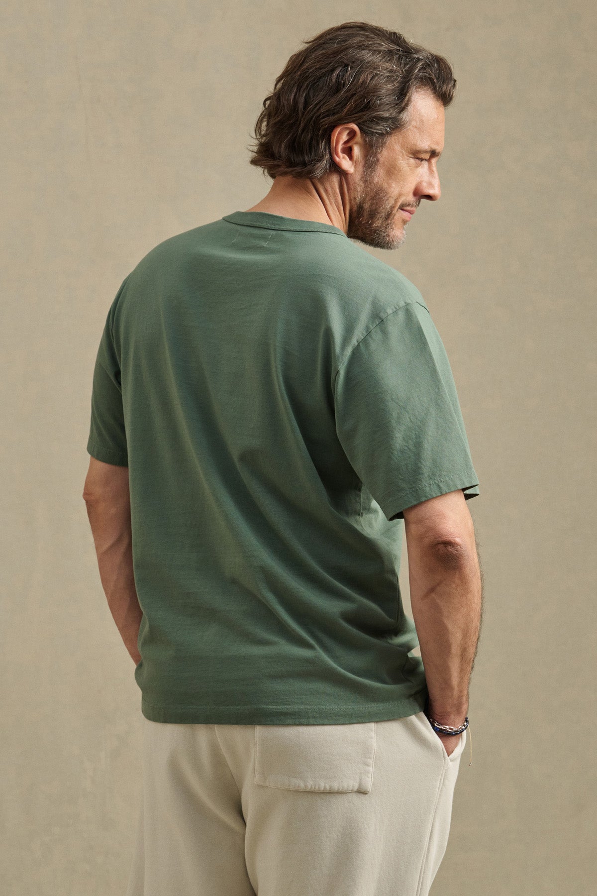 A man stands with his back turned, wearing the WELLS TEE by Velvet by Graham & Spencer—a relaxed fit green short-sleeve Pima cotton jersey t-shirt paired with beige pants, set against a plain neutral background. Perfect for all-day comfort.-41480101494977