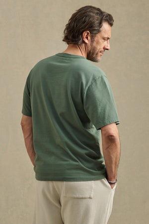 A man stands with his back turned, wearing the WELLS TEE by Velvet by Graham & Spencer—a relaxed fit green short-sleeve Pima cotton jersey t-shirt paired with beige pants, set against a plain neutral background. Perfect for all-day comfort.