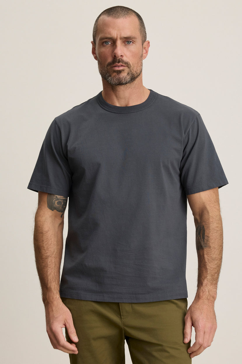 A man with short hair and a beard is wearing the WELLS TEE by Velvet by Graham & Spencer, a plain dark gray cotton jersey crewneck, paired with olive green pants against a neutral background.