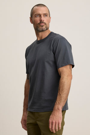 A man with a short beard and close-cropped hair wears the WELLS TEE by Velvet by Graham & Spencer in dark gray cotton jersey with a crew neckline and olive green pants, standing against a plain light background.