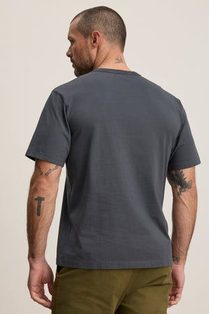 A man with short hair and arm and neck tattoos wears the WELLS TEE by Velvet by Graham & Spencer—a relaxed fit, dark gray cotton jersey crewneck—paired with olive pants, shown from the back against a neutral background.