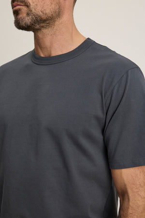 A man with a short beard is wearing the WELLS TEE by Velvet by Graham & Spencer, a relaxed fit dark gray crew neck t-shirt. The image is cropped from his shoulders to just above his mouth.