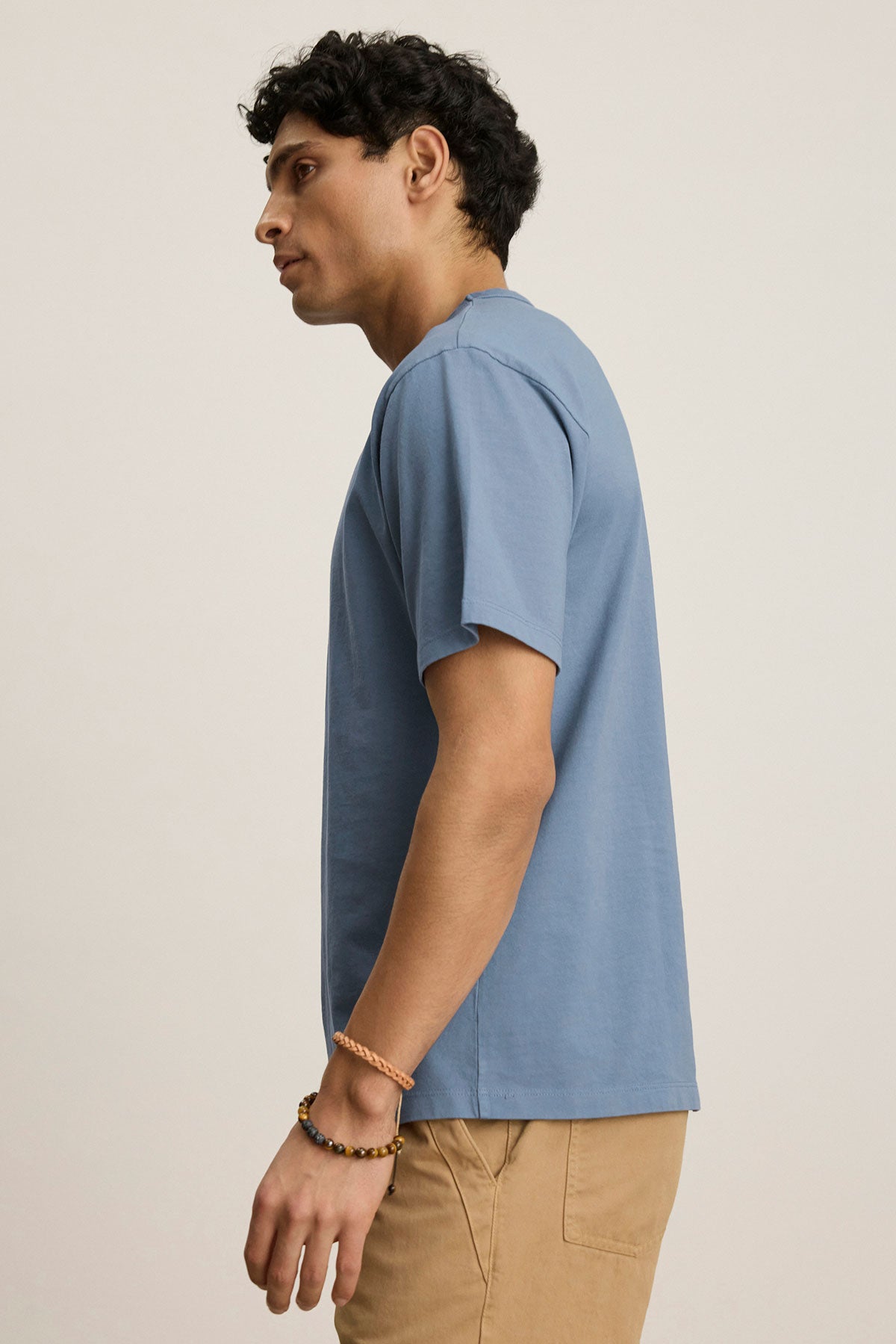 A man stands in profile wearing the WELLS TEE by Velvet by Graham & Spencer, a relaxed fit blue cotton jersey crewneck, paired with beige pants against a plain, light background.-39808332824769