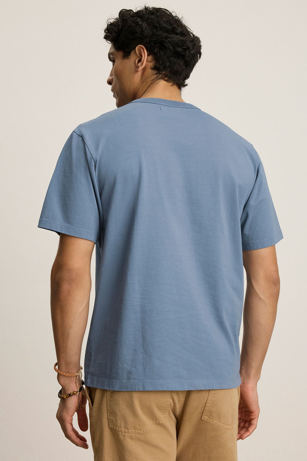 A man faces away from the camera, wearing the WELLS TEE by Velvet by Graham & Spencer—a light blue cotton jersey crewneck—paired with tan pants against a neutral background.-39808332857537