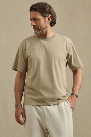 A man with short brown hair and a beard wears the Velvet by Graham & Spencer WELLS TEE, a relaxed fit beige Pima cotton jersey top, paired with light pants. He stands against a plain tan background, looking to his left. Ideal for all-day comfort.