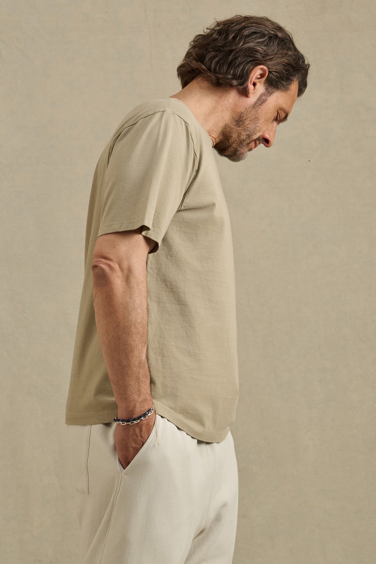 A man with dark hair and a beard stands in profile, wearing the WELLS TEE by Velvet by Graham & Spencer—crafted from soft Pima cotton jersey—for all-day comfort, paired with light pants against a neutral background.