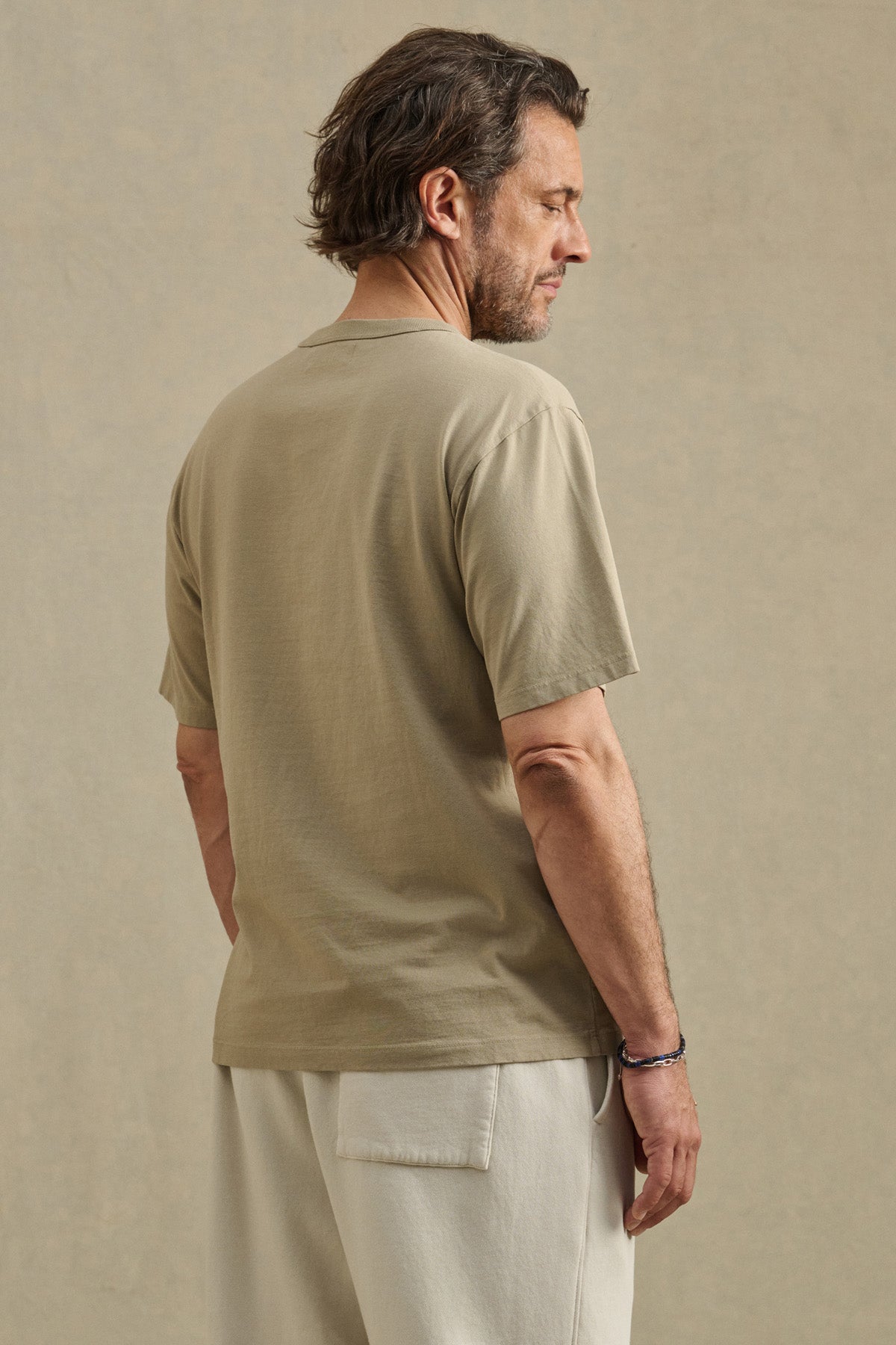 A man with brown hair is standing and facing away, wearing the WELLS TEE by Velvet by Graham & Spencer—a beige, relaxed fit short-sleeve crew neck t-shirt—paired with light pants against a plain background.