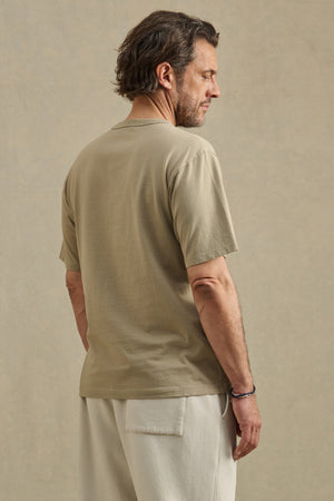 A man with brown hair is standing and facing away, wearing the WELLS TEE by Velvet by Graham & Spencer—a beige, relaxed fit short-sleeve crew neck t-shirt—paired with light pants against a plain background.