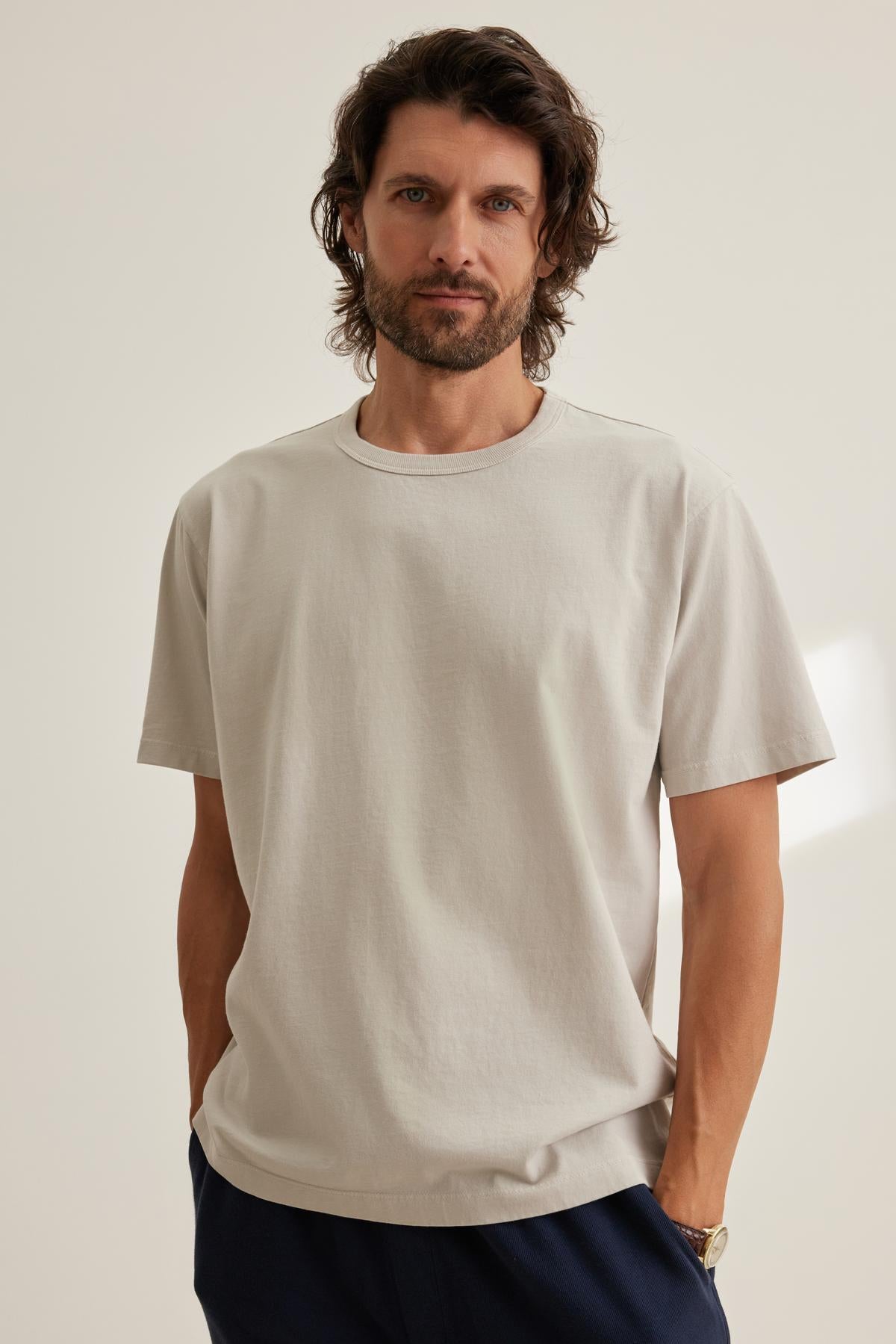   A man with medium-length hair and a beard wears the Velvet by Graham & Spencer WELLS TEE, a plain light beige t-shirt, paired with dark pants, as he stands with hands in pockets against an off-white background. 