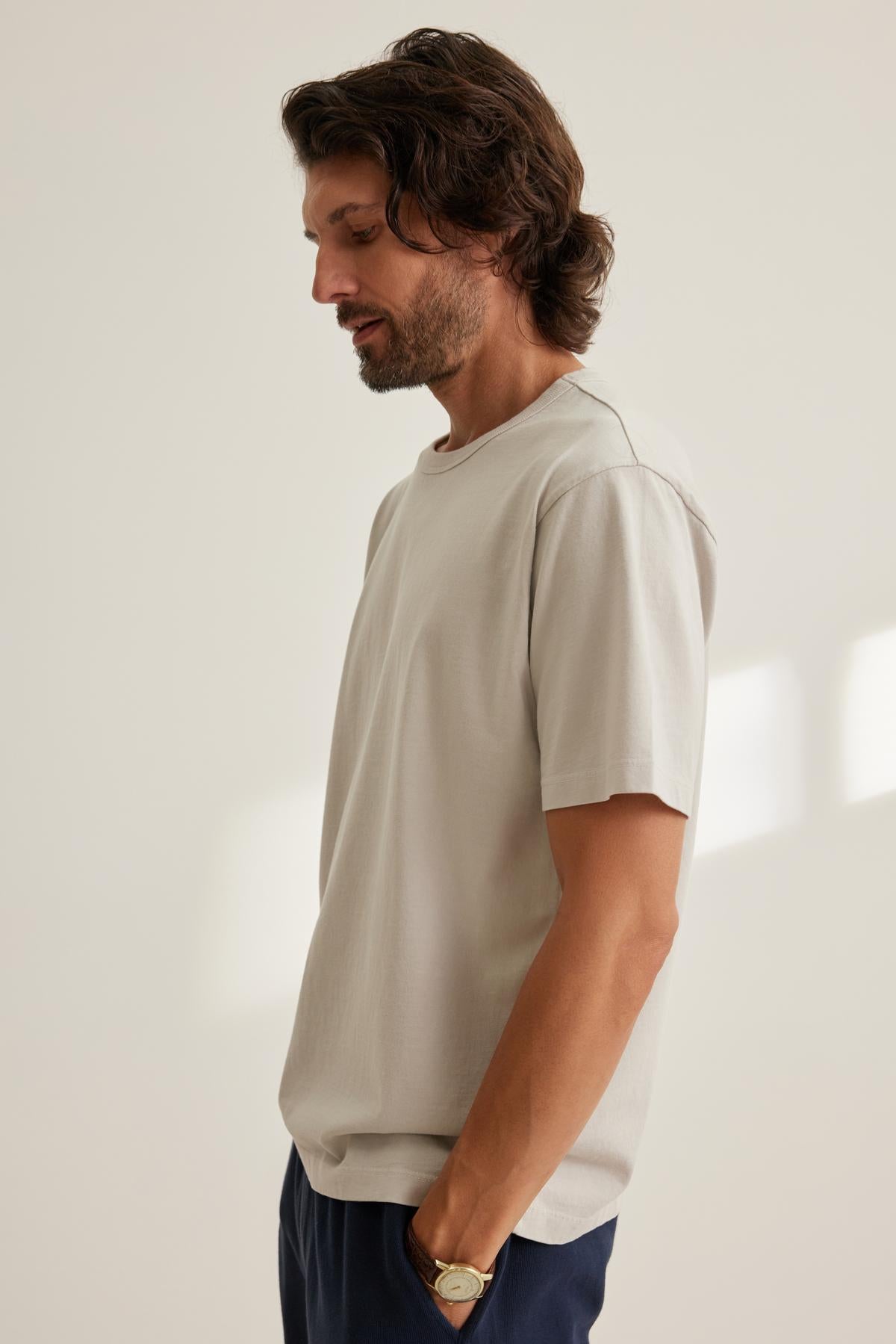 A man with shoulder-length hair and a beard stands in profile, wearing a Velvet by Graham & Spencer WELLS TEE, white in color, along with dark pants, and has his hands in his pockets.-38643375734977