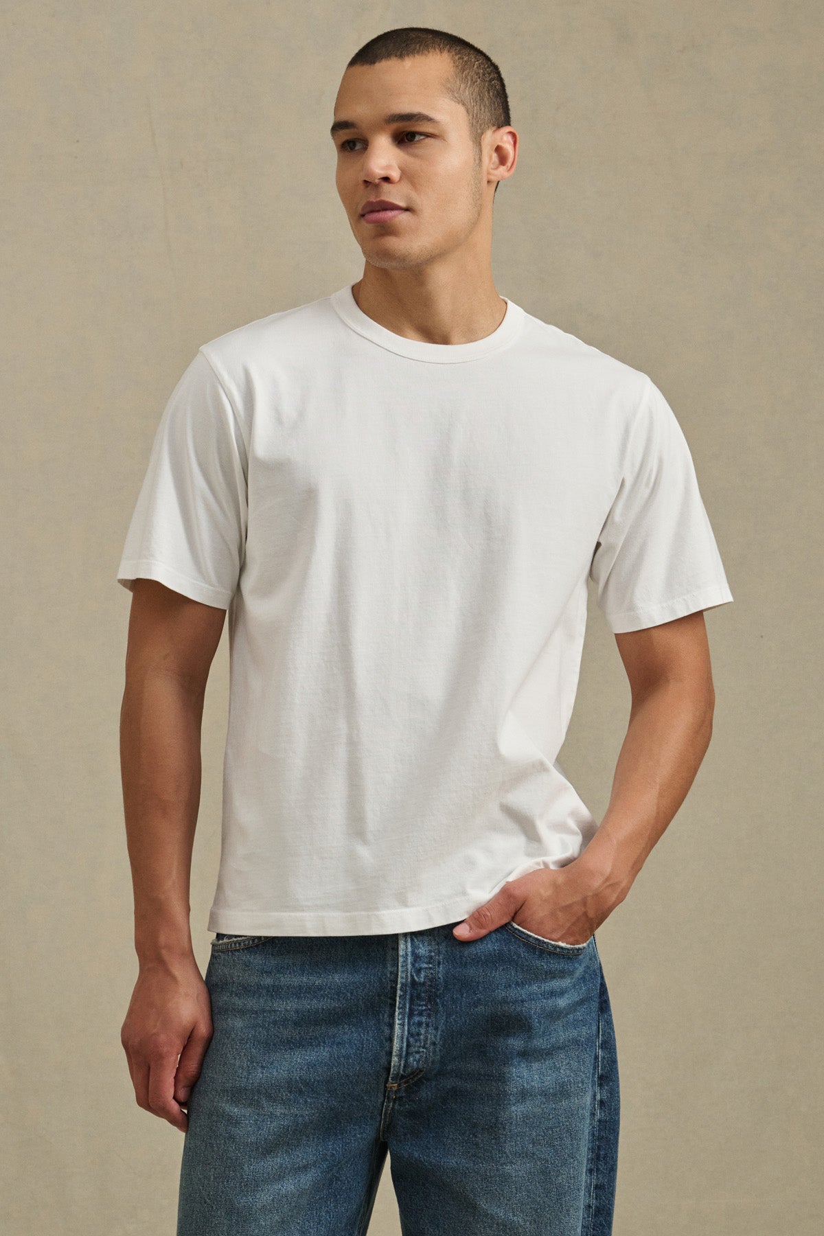 A man wears the WELLS TEE by Velvet by Graham & Spencer—a plain white Pima cotton jersey crew neck short-sleeve t-shirt—with blue jeans, standing against a neutral background and looking slightly to the side.-41548224889025