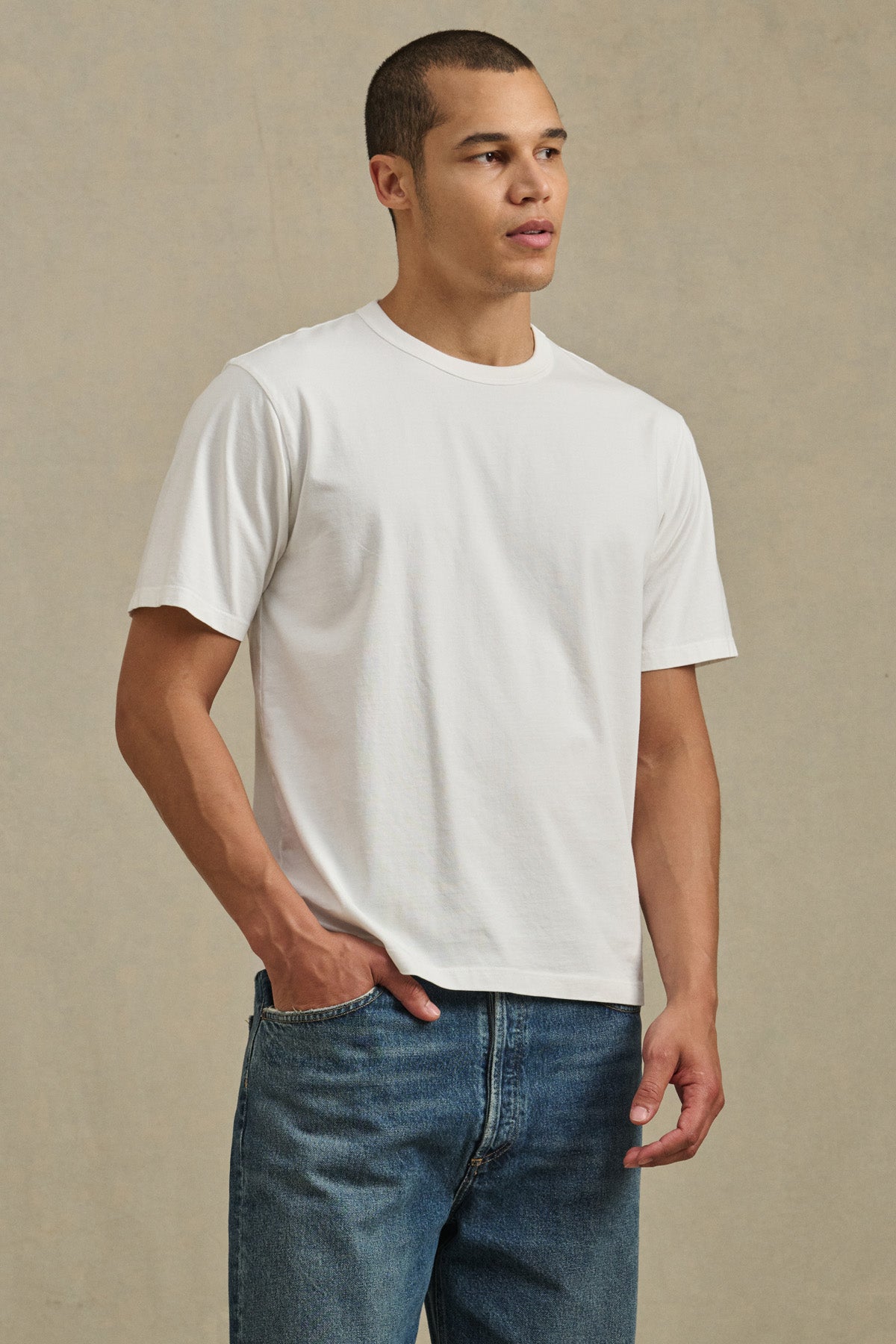 A person stands against a neutral background, wearing the WELLS TEE by Velvet by Graham & Spencer—a relaxed fit plain white short-sleeve crewneck—paired with blue jeans, one hand in their pocket.-41548224921793
