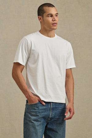 A person stands against a neutral background, wearing the WELLS TEE by Velvet by Graham & Spencer—a relaxed fit plain white short-sleeve crewneck—paired with blue jeans, one hand in their pocket.