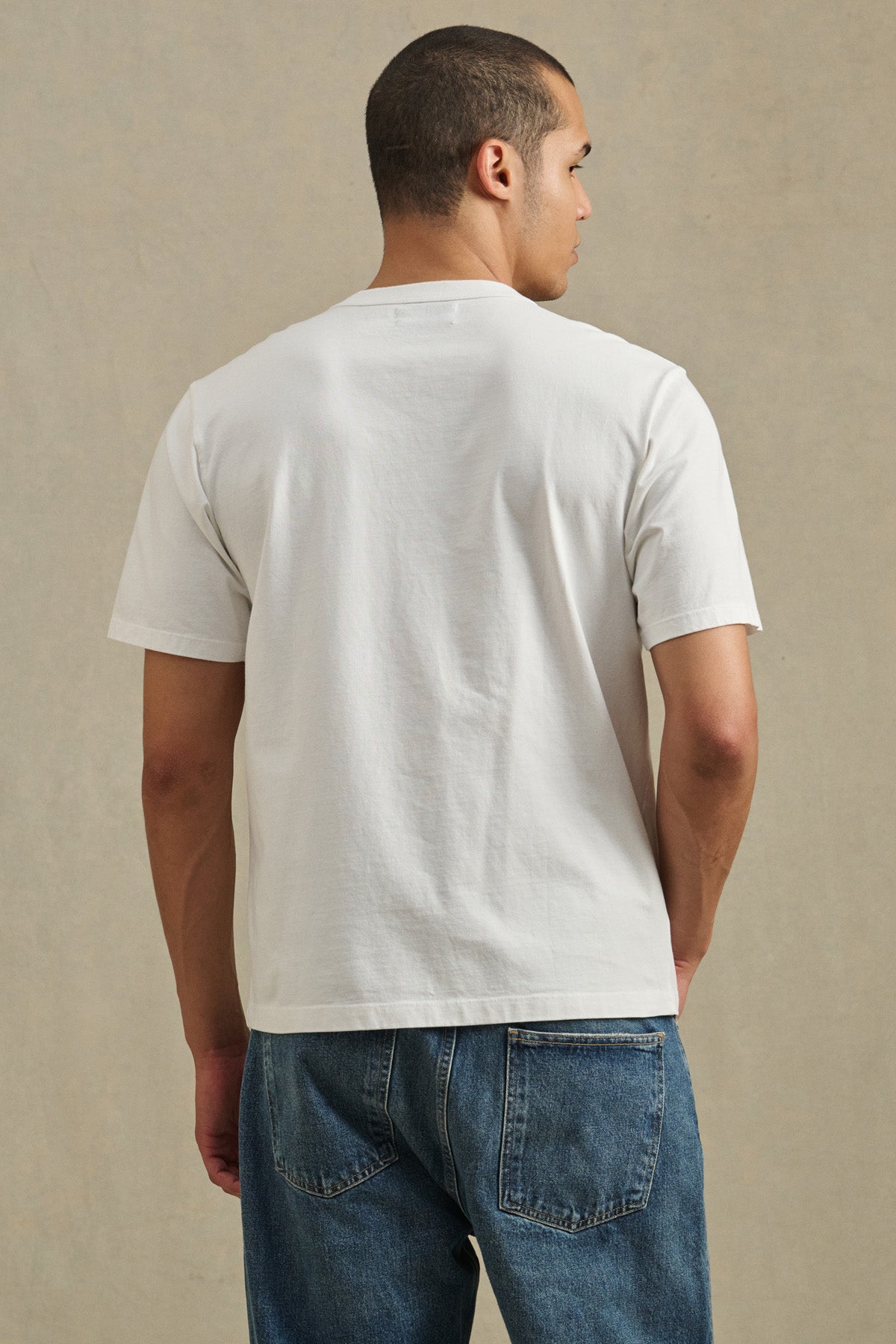 A man faces away from the camera, wearing the WELLS TEE by Velvet by Graham & Spencer—a plain white Pima cotton crewneck t-shirt—paired with blue jeans against a neutral background.-41548224987329