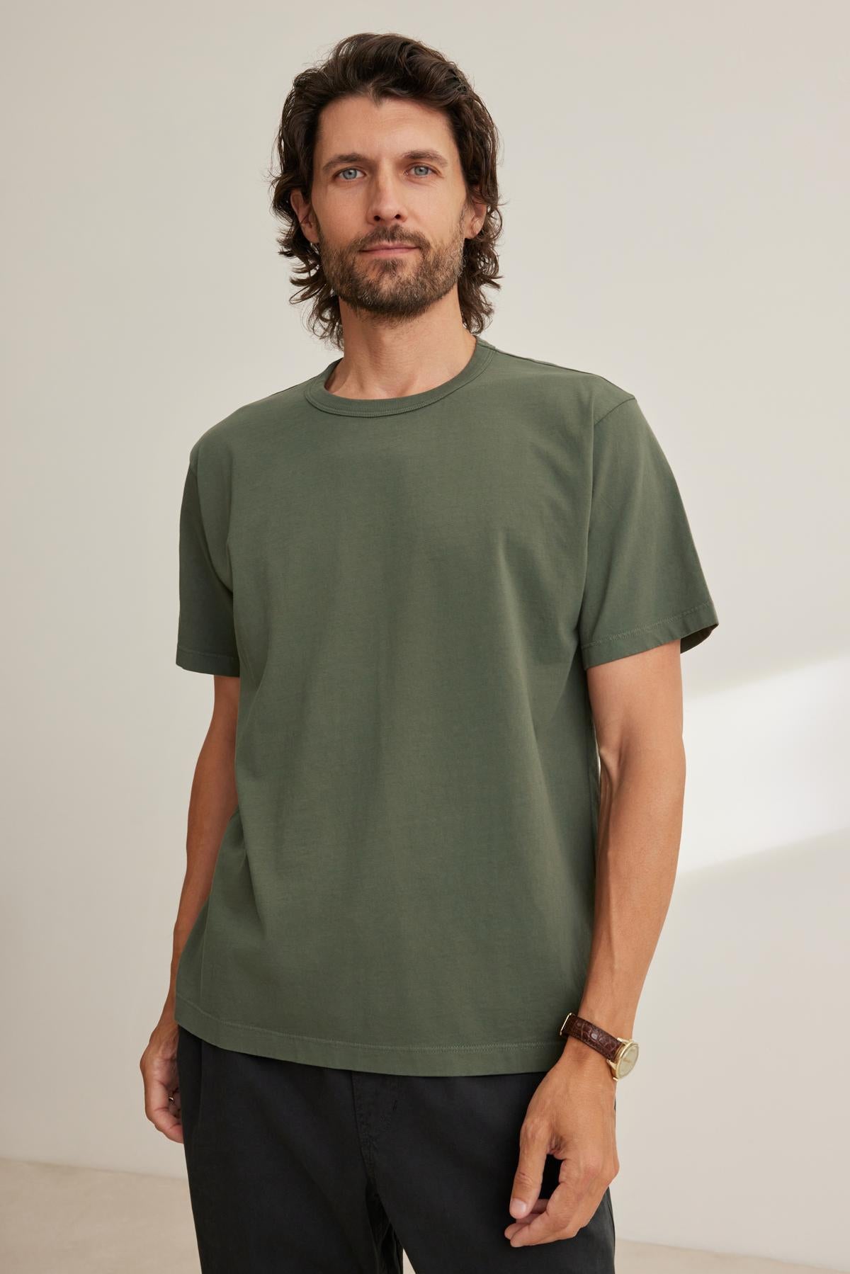 A man with dark hair and a beard wears the WELLS TEE by Velvet by Graham & Spencer, paired with black pants, standing against a plain beige background.-38643375636673