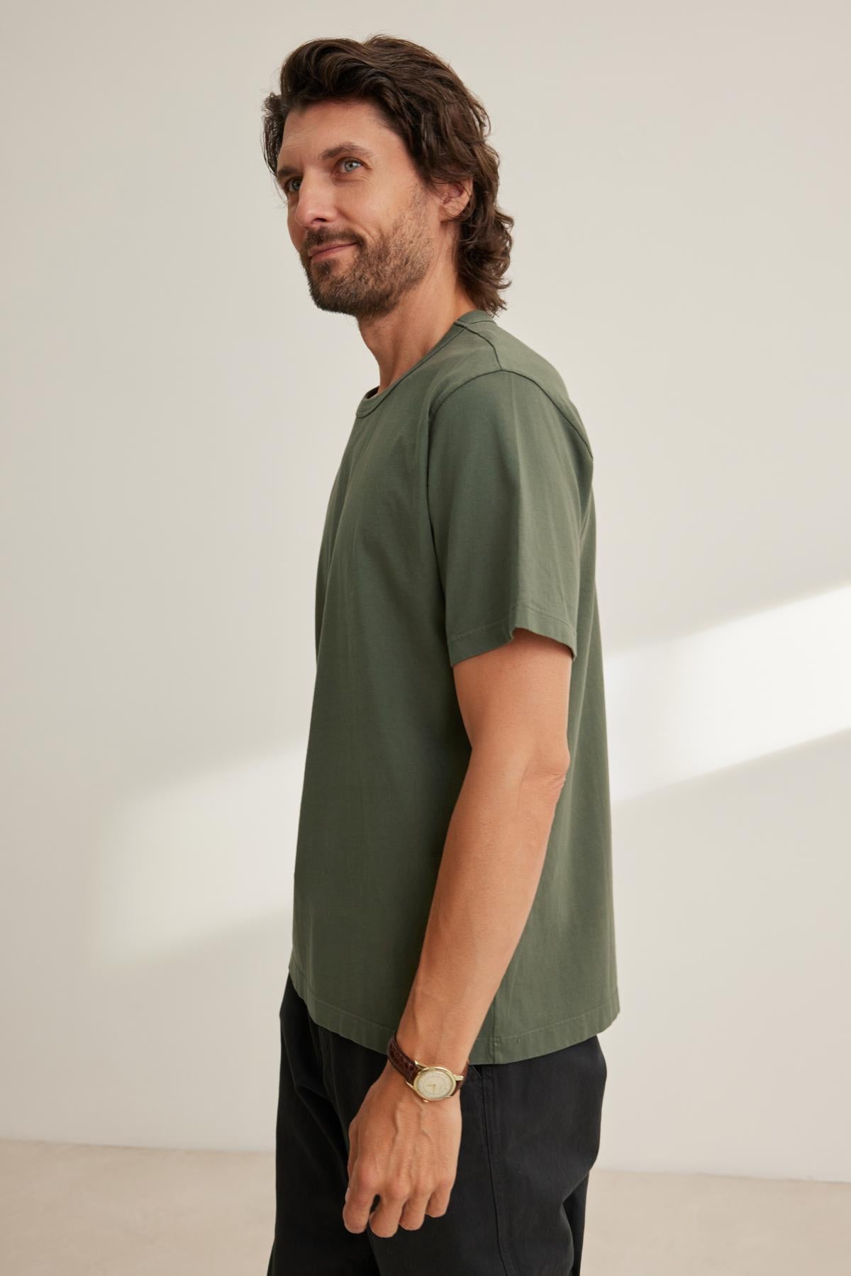   A man with a beard wearing Velvet by Graham & Spencer's WELLS TEE and dark pants stands sideways in a well-lit room. 