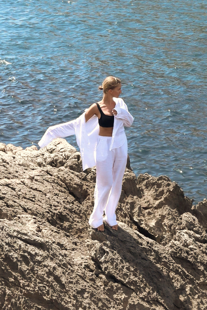   A woman in a white PICO PANT by Velvet by Jenny Graham top and pants standing on a rocky beach. 