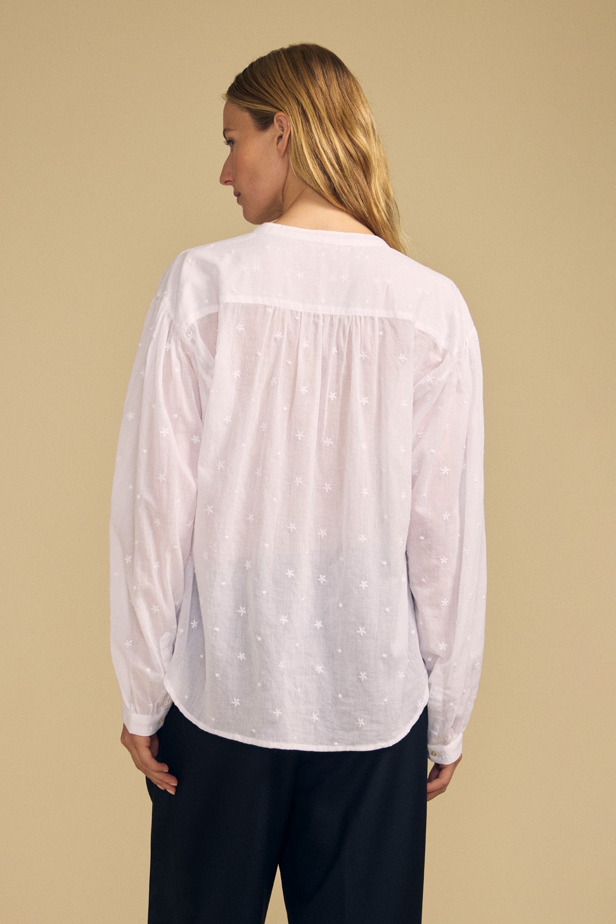 A person with long hair, seen from behind, wears the JULIANNE BLOUSE by Velvet by Graham & Spencer—a white long-sleeve blouse featuring subtle embroidered patterns—paired with black pants against a beige background.-41826785231041