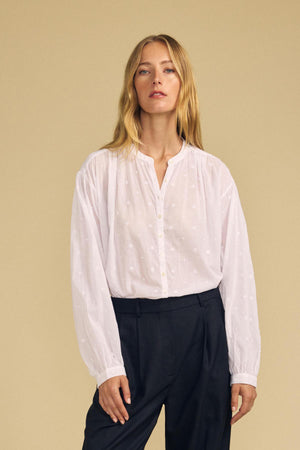 A woman wearing the Velvet by Graham & Spencer JULIANNE BLOUSE—a loose white button-up with subtle embroidery—paired with black high-waisted trousers, standing against a plain beige background.
