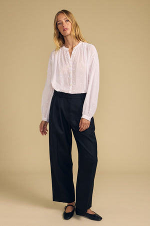 A person stands against a plain beige background, wearing the Velvet by Graham & Spencer JULIANNE BLOUSE in white, paired with black wide-leg pants and black flats.