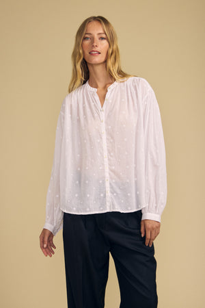 A person wearing the JULIANNE BLOUSE by Velvet by Graham & Spencer—a loose, white, long-sleeve button-up blouse with subtle embroidery—paired with black pants stands against a beige background.