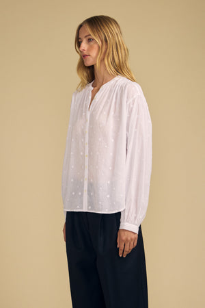 A person stands against a beige background wearing the JULIANNE BLOUSE by Velvet by Graham & Spencer— a white long-sleeve blouse with a subtle dotted pattern—paired with loose black trousers.