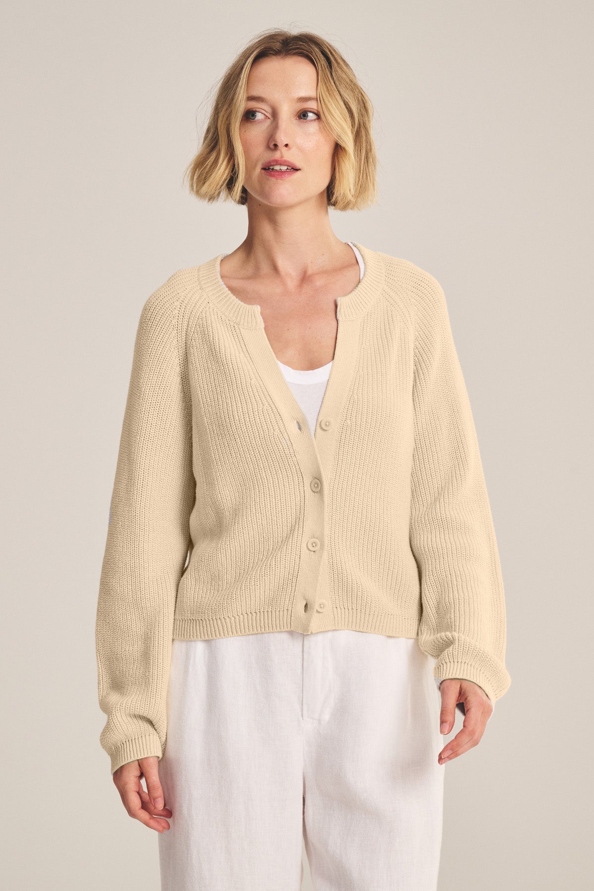 A person wears the MIRAE CARDIGAN by Velvet by Graham & Spencer over a white top and white pants, standing against a plain, light-colored background.