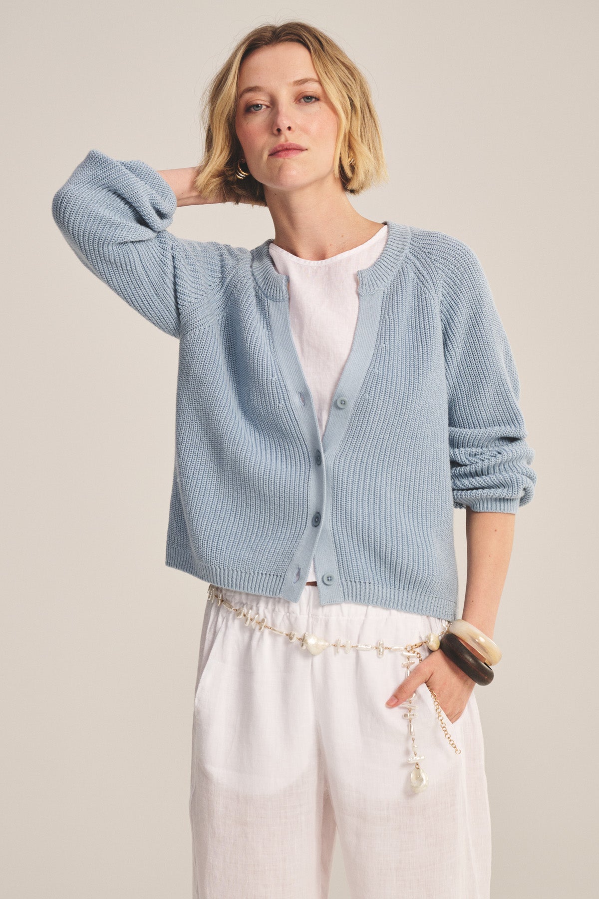 A woman wears the MIRAE CARDIGAN by Velvet by Graham & Spencer in light blue over a white outfit, accessorized with a beaded belt and bracelets, standing with one hand behind her head.-40909904281793