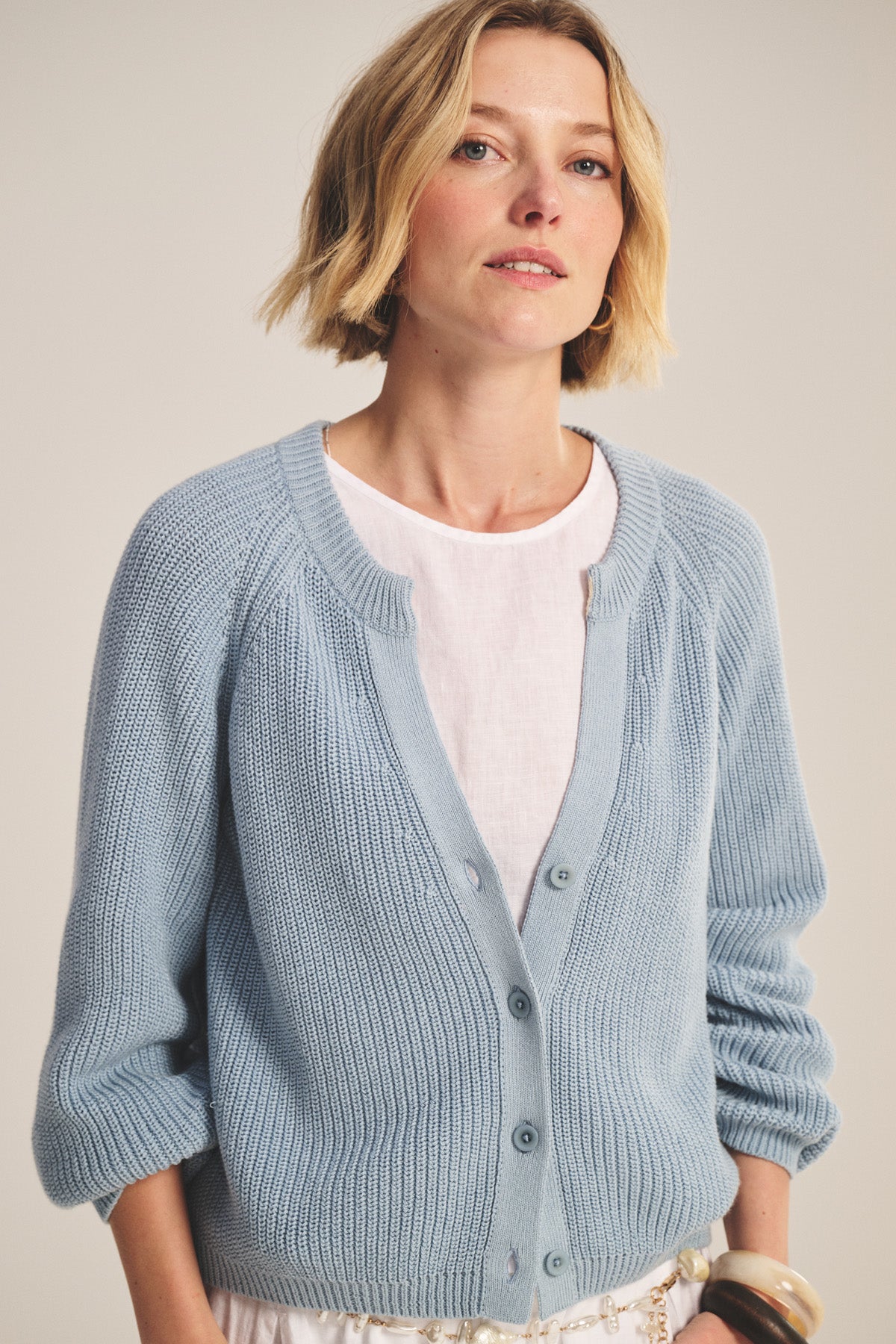 A woman with short blonde hair models the MIRAE CARDIGAN by Velvet by Graham & Spencer, a light blue textured knit, worn over a white top against a plain background.-40909904249025