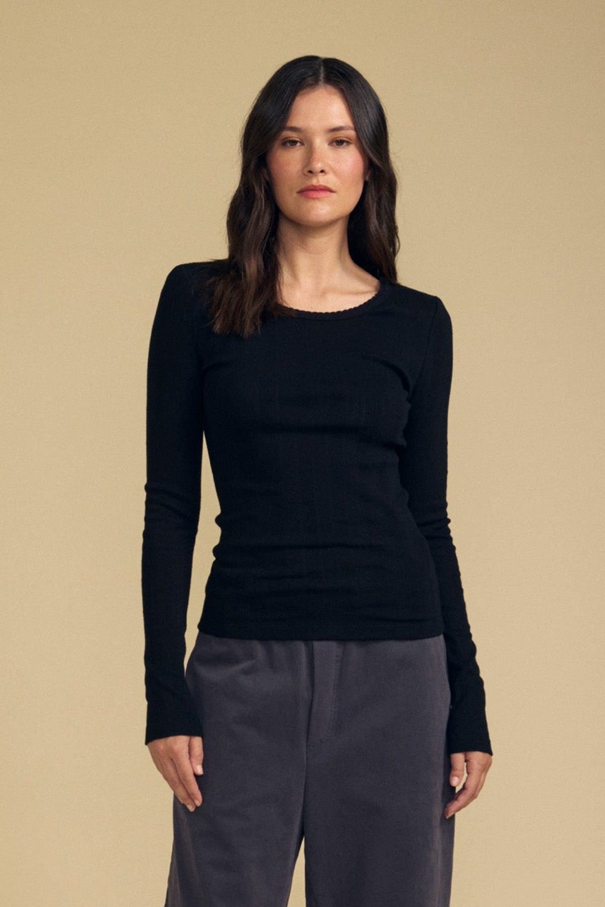 A woman with long dark hair stands facing forward against a plain beige background, wearing the Velvet by Graham & Spencer MIRIAM TEE—a fitted black cotton long sleeve top with a scalloped crew neckline—paired with loose gray pants.