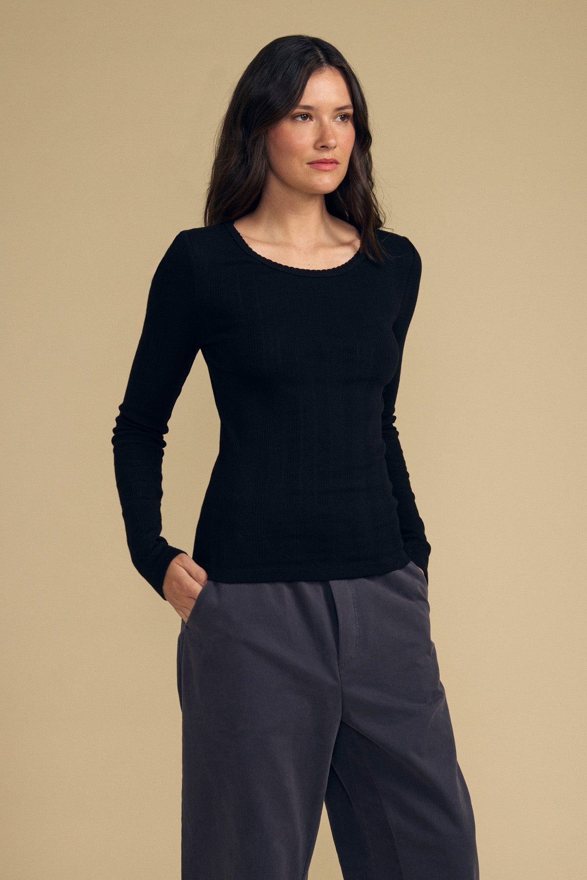 A person stands against a beige background, wearing the MIRIAM TEE by Velvet by Graham & Spencer—a cotton long sleeve black top with a scalloped crew neckline—paired with gray pants, hands in pockets, facing slightly to the right.-41838213693633