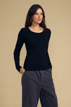 A person stands against a beige background, wearing the MIRIAM TEE by Velvet by Graham & Spencer—a cotton long sleeve black top with a scalloped crew neckline—paired with gray pants, hands in pockets, facing slightly to the right.