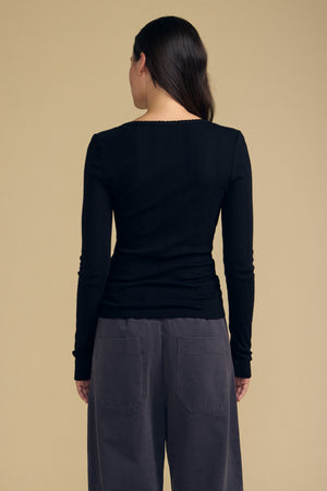 A woman stands with her back to the camera against a beige background, wearing the MIRIAM TEE by Velvet by Graham & Spencer—a fitted black cotton long sleeve top with a scalloped crew neckline—paired with dark gray high-waisted pants.