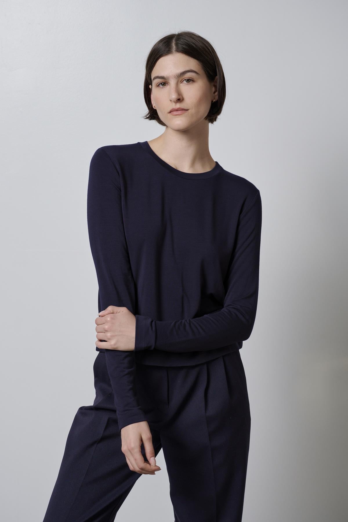   A person with short dark hair stands against a plain background, wearing the Velvet by Jenny Graham PACIFICA TEE and matching pants that feature a long-sleeve navy rib knit design, offering a slimmer fit and added stretch, with their arms crossed. 