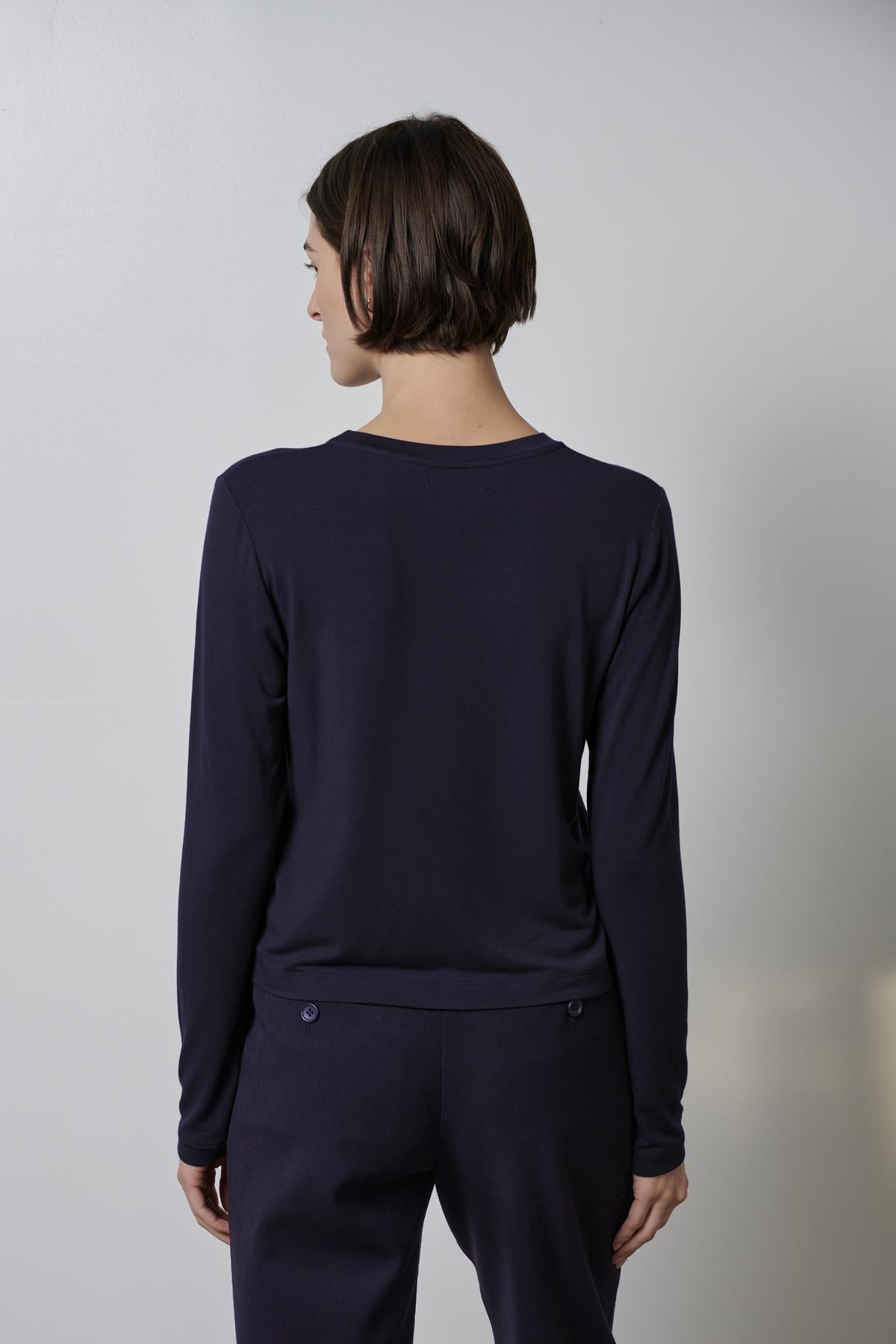   A person with short hair is standing with their back to the camera, wearing the PACIFICA TEE by Velvet by Jenny Graham and navy pants against a plain light-colored background. 