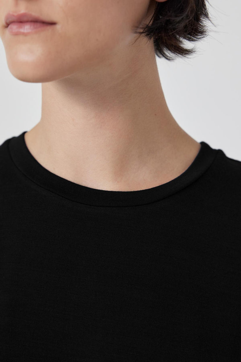 Close-up view of a person wearing a black SOLANA TEE by Velvet by Jenny Graham with short hair, partially showing their face against a plain, light background. The shirt's tailored finish highlights the soft modal jersey fabric.