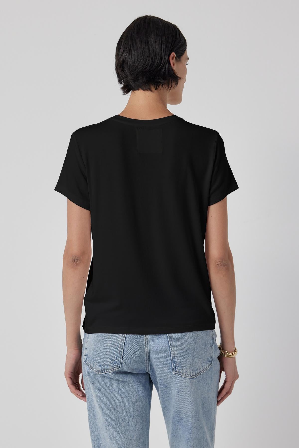 A person with short dark hair, wearing a tailored finish black SOLANA TEE by Velvet by Jenny Graham and light blue jeans, is standing with their back to the camera against a plain background.-36463664070849