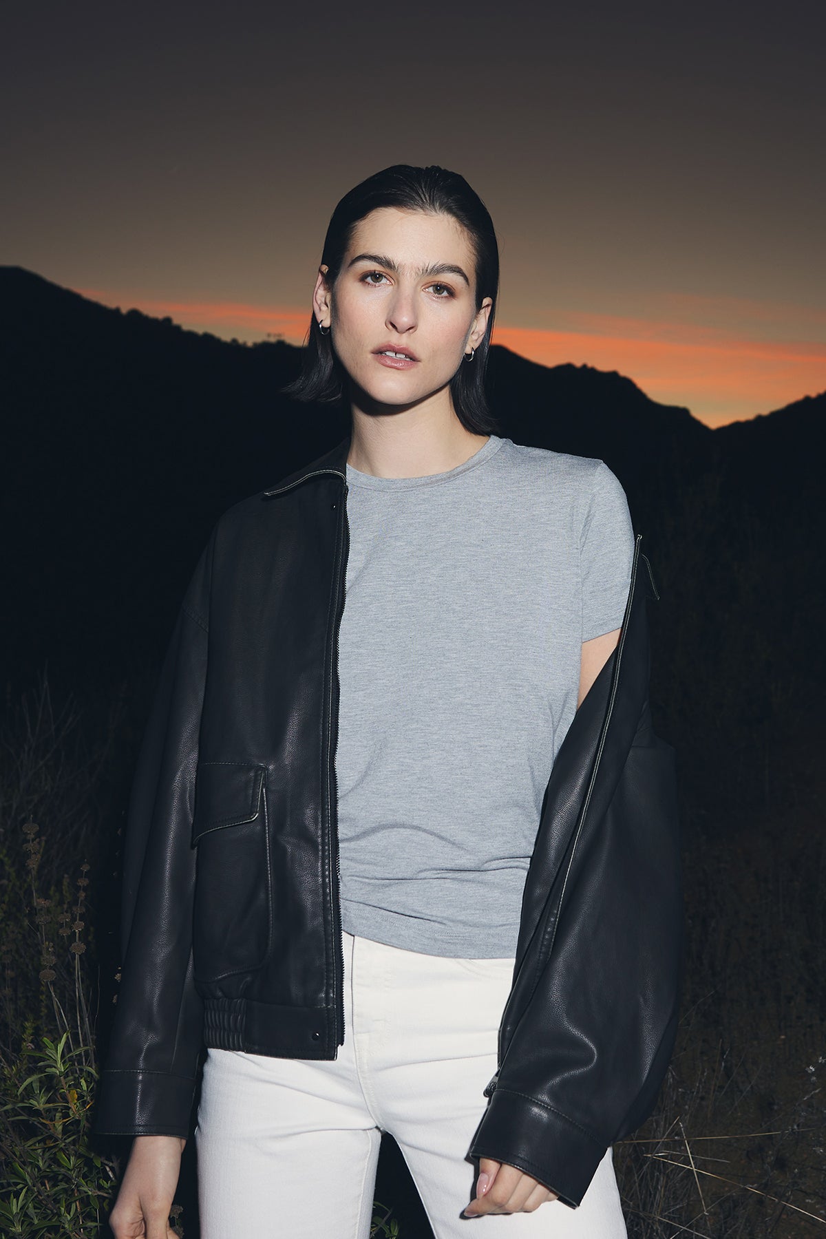   A person with short dark hair stands outdoors at dusk, wearing a light gray SOLANA TEE from Velvet by Jenny Graham, white pants, and a partially worn black leather jacket. Hills are visible in the background against a sunset sky. 