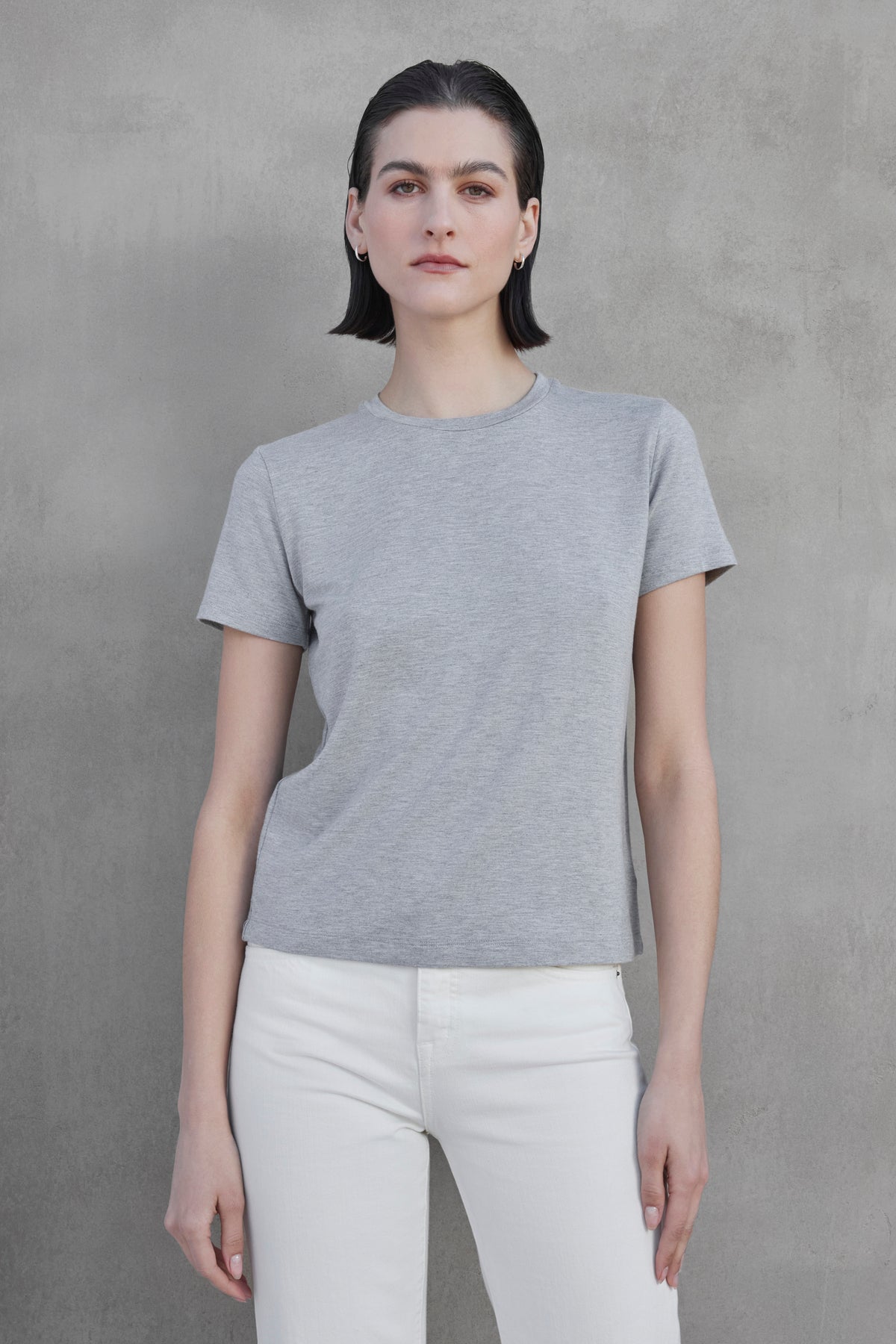 A person with short dark hair is wearing a gray SOLANA TEE by Velvet by Jenny Graham and white pants, standing against a bare, textured gray wall.-37659920728257